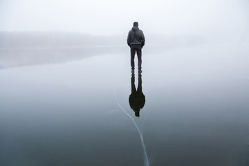Young adult man standing alone on cracked dark ice surface. Mist over frozen lake in winter. Foggy...