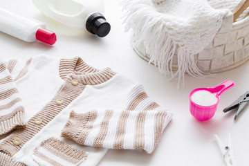 White and light brown, warm child sweater on table. Different washing detergents. Pink cup or scoop...