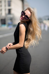 Adorable smiling blonde woman wearing black dress and sunglasses, walking down the sunny street