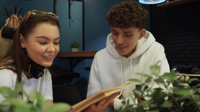 Girl reads her boyfriend a funny excerpt from fiction book and they laugh and smile sitting in coffee house