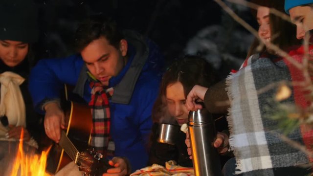Group of friends having a good time by the fire in the woods. Having a snack. A young man tuning the guitar