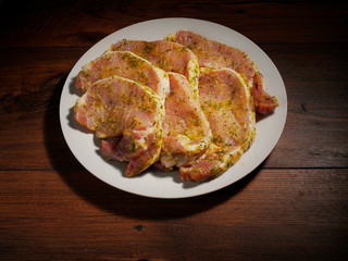 Raw marinated pork chops on a white plate wooden table, high contrast lighting.