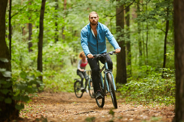 Fototapeta na wymiar Active guy in sportswear cycling along forest path on summer day with his girlfriend moving after him