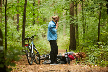 Young female cyclist with broken leg lying on forest path while her boyfriend calling for help on...