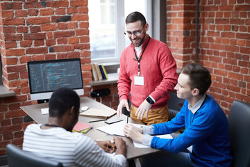 Young successful coach standing by table and discussing new ideas with two specialists at training