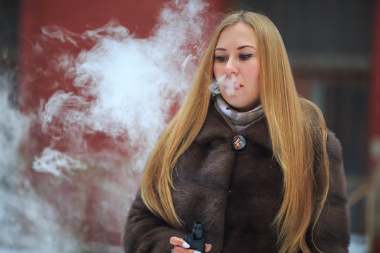 Vape lady. Young pretty girl blonde in a fur coat smokes an electronic cigarette in a vintage yard outdoors in the winter.