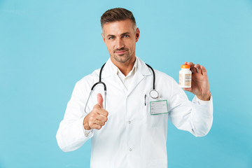 Mature man doctor posing isolated over blue wall background holding pills vitamins showing thumbs up.