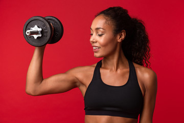 Image of beautiful african american woman in black sportswear lifting dumbbell, isolated over red background