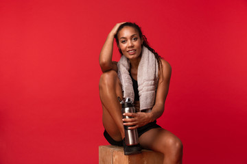 Obraz premium Portrait of joyous african american woman in black sportswear sitting on box with towel and water bottle, isolated over red background