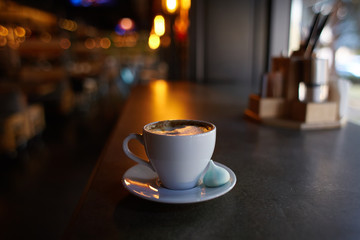 Obraz premium Cup of americano coffee in a white cup with a marshmallow on a saucer, shallow focus, dark background, bulb lights of caffeine