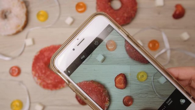 Mobile photography on smartphone camera of donuts, sweetmeats candy and strawberry on wooden background. Coconut and berry homemade glazed and filled donuts. Making photo of colorful sugar and sweets.
