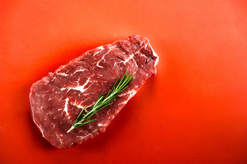Raw beef steak on a red surface. Steak decorated with a sprig of rosemary. Close-up. View from...