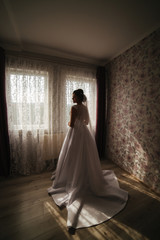 Charming bride stand in front of window. Girl stand by the window at her room