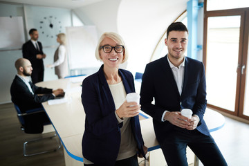 Two happy elegant colleagues with drinks standing by table in boardroom during coffee break