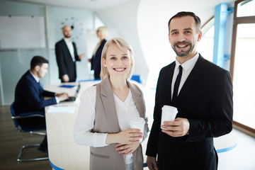 Two elegant cheerful colleagues with glasses of coffee looking at you in boardroom at break