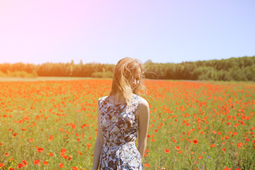 Naklejka na ściany i meble Beautiful girl is standing on summer field full of red poppy flowers in the grass. Sunny day with green lawn background. Happy woman in rustic dress is smiling. The wind in her hair.