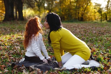 Autumn vibes, family portrait. Charming mom and her red hair daughter have fun sitting on the fallen leaves in the park full of evening sun