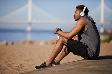 Serious thoughtful young black man in gray sleeveless hoodie sitting on ground and listening to...