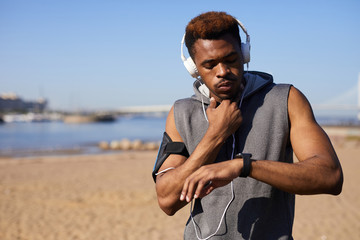 Serious pensive young black man in wired headphones connect to smartphone in armband touching neck...