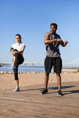 Content confident motivated young multi-ethnic couple in sportswear stretching on beach before jogging in summer