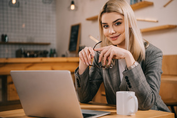 selective focus of smiling woman looking at camera near laptop and holding glasses in cafe
