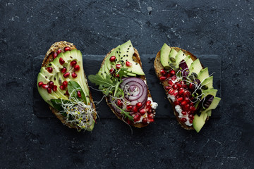 Healthy avocado toasts with avocado slices, pomegranate seeds and sprouts on dark background.