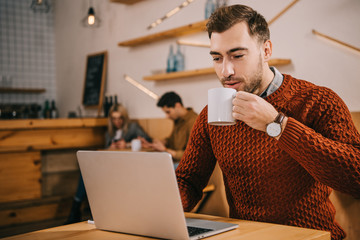 handsome man drinking coffee and looking at laptop in cafe