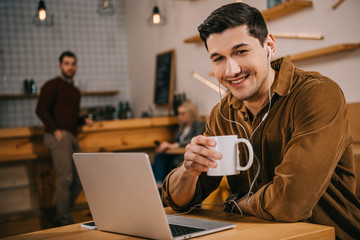 handsome man in earphones holding cup of coffee near laptop in cafe