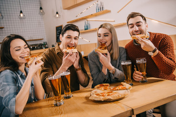 happy friends eating pizza near glasses of beer in bar