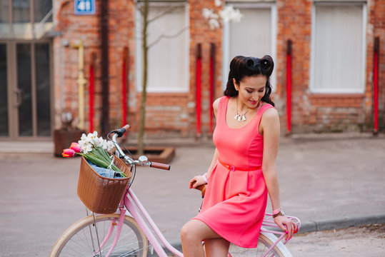 Young attractive woman in pink dress and her a bicycle in the city street with flowers during an exotic summer trip on vacation. Sunny day outdoors.