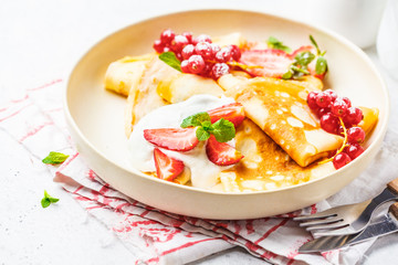 Homemade thin crepes served with curd cream, currants, strawberries and powdered sugar in white plate.