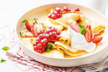 Homemade thin crepes served with curd cream, currants, strawberries and powdered sugar in white plate.