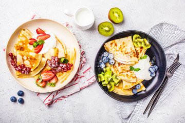 Homemade thin crepes served with curd cream, fruits and berries in black and white plates, top view.