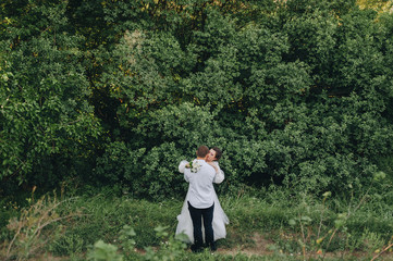 Beautiful newlyweds hugging in nature, against the background of green foliage and trees. Young bridegroom in a white shirt hugs a beautiful bride in sunny weather. Wedding photography.