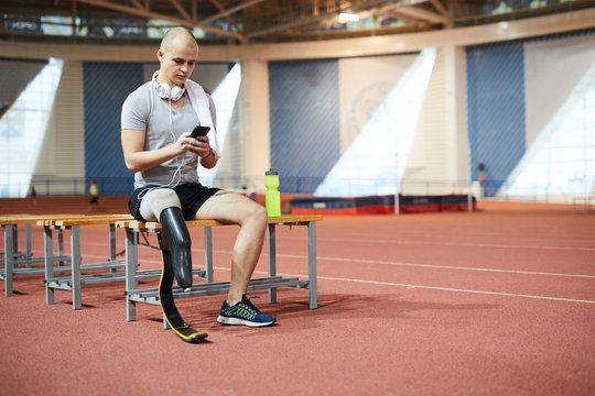 Young paralympic champion or competitor with headphones sitting on bench while scrolling in smartphone for playlist at break