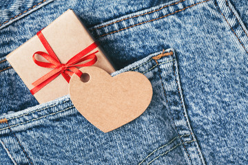 Kraft gift box with red bow and tag in shape of heart sticking out of blue jeans pocket. Valentine's day card.