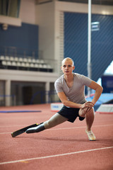 Fototapeta premium Young man in activewear doing stretching exercise for legs on racetrack on stadium while preparing for paralympic games