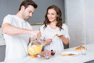 Obraz na płótnie Canvas girlfriend looking at handsome man holding jug with orange juice in kitchen
