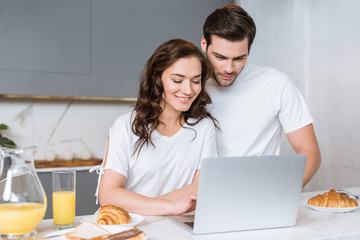 happy woman looking at laptop near handsome boyfriend in kitchen