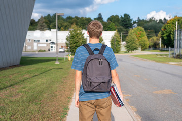 Male teenager with his back turned to the camera and walking towards a school. He is wearing a backpack and carrying some binders.