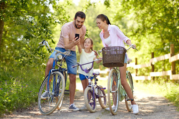 family, leisure and technology concept - happy mother, father and little daughter with smartphone and bicycles in summer park