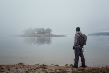 Fototapeta na wymiar Winter mountain hiking Rear view of a man with a backpack hiking in the winter Rhodopi Mountains in Bulgaria and enjoys the view of the lake and covered by fog islet