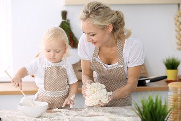 Obraz na płótnie Canvas Little girl and her blonde mom in beige aprons playing and laughing while kneading the dough in kitchen. Homemade pastry for bread, pizza or bake cookies. Family fun and cooking concept
