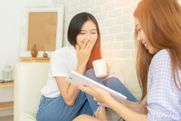 Obraz na płótnie Canvas Happy female friends holding coffee mugs while discussing at table in living room relax time.Asian beauty woman smile and sititing sofa .she shopping online .