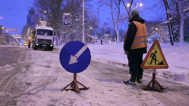 Snowblower clear freezing winter road with snow and ice. Snow-plow remove snow from the city street. Worker install warning road signs. Cleaning snowy frozen roads.