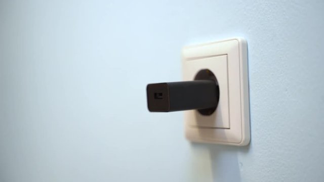 Woman hand plug black phone charger adapter into wall socket.