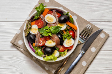 Top view, flat lay. Traditional niçoise (nicoise) salad in a white plate on a polka dot napkin. Near cutlery. White background.
