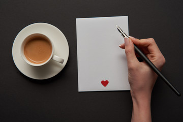 cropped view of woman writing on empty greeting card with red heart sign near cup of coffee