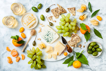 Cheese platter with different cheeses, grapes, nuts, honey. Appetizers table with antipasti snacks. Cheese variety board over white marble background. Top view, flat lay