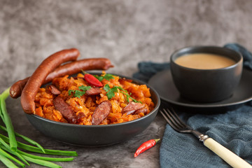 Cabbage stew in bowl with smoked sausagesand cup of coffee on grey background. Side view